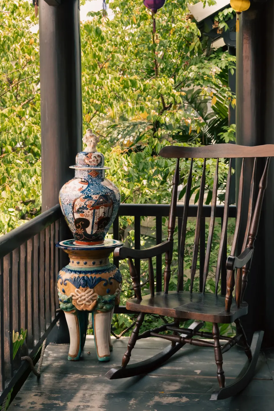 A rocking chair and a painted jar on a shaded wooden veranda among green leaves.