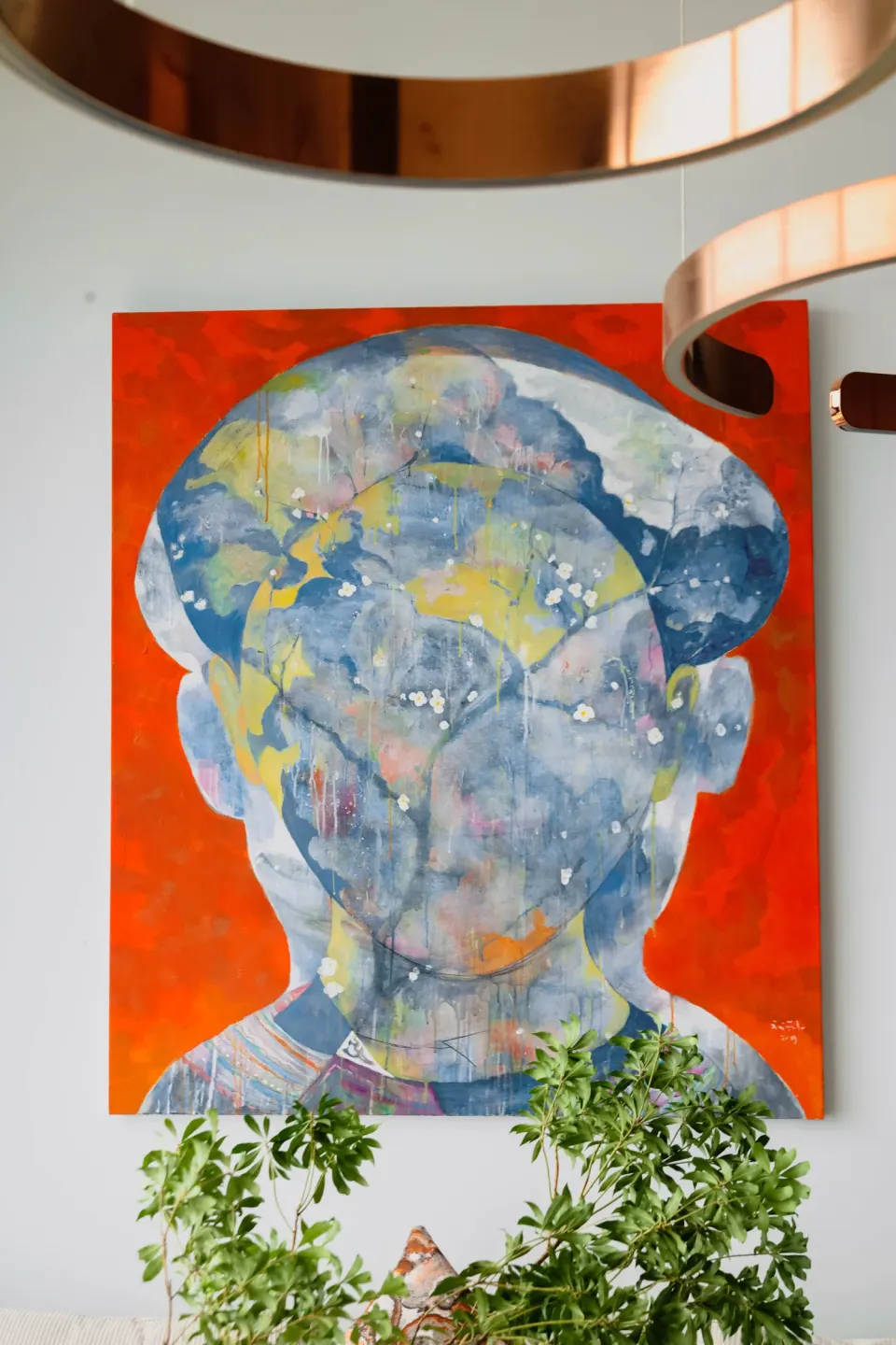 A large orange abstract portrait painting on a white wall under copper ring lights.