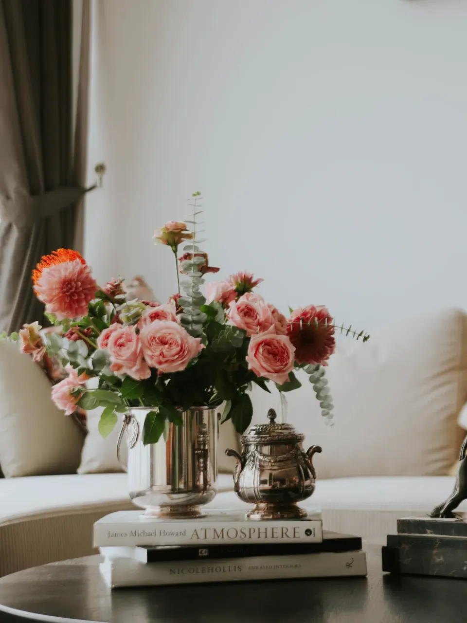 Pink roses and dahlias in a silver vase on a coffee table beside a bronze figure.