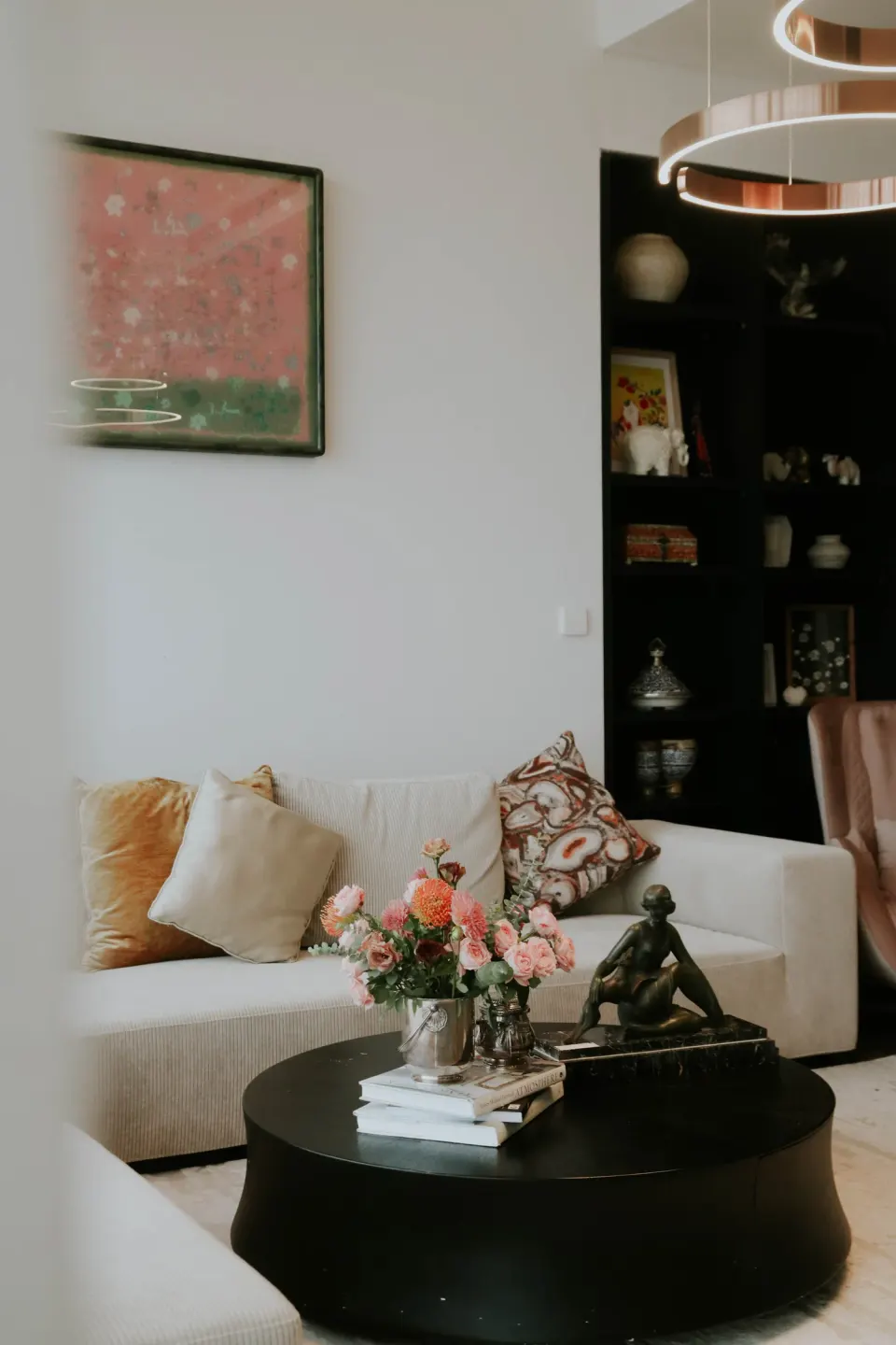 A pink painting above a sofa, a bouquet and a bronze figure on the round table.