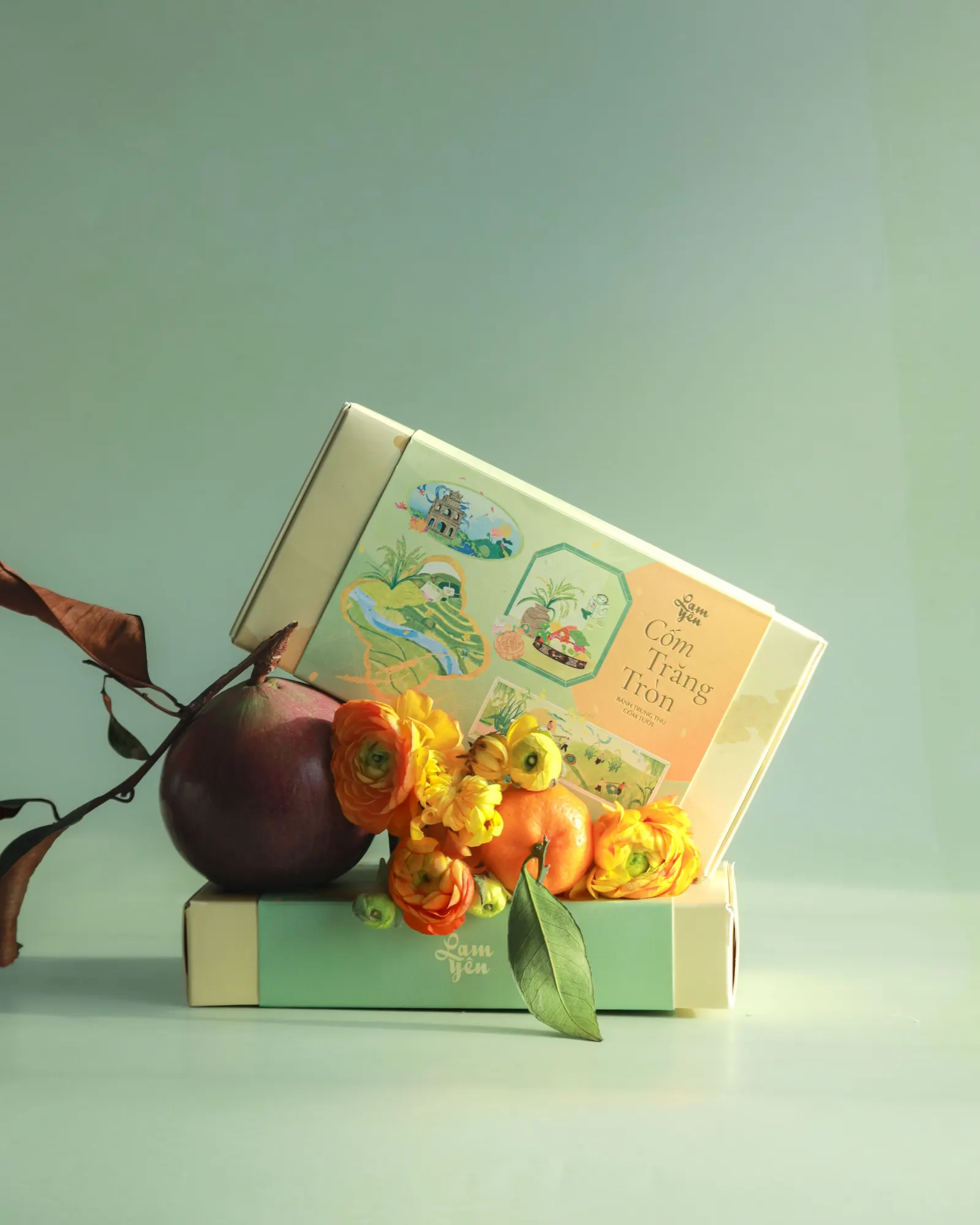 A pale green Lam Yên gift box tilted open on a box, with orange ranunculus, a persimmon and a dried branch, against a soft green backdrop.
