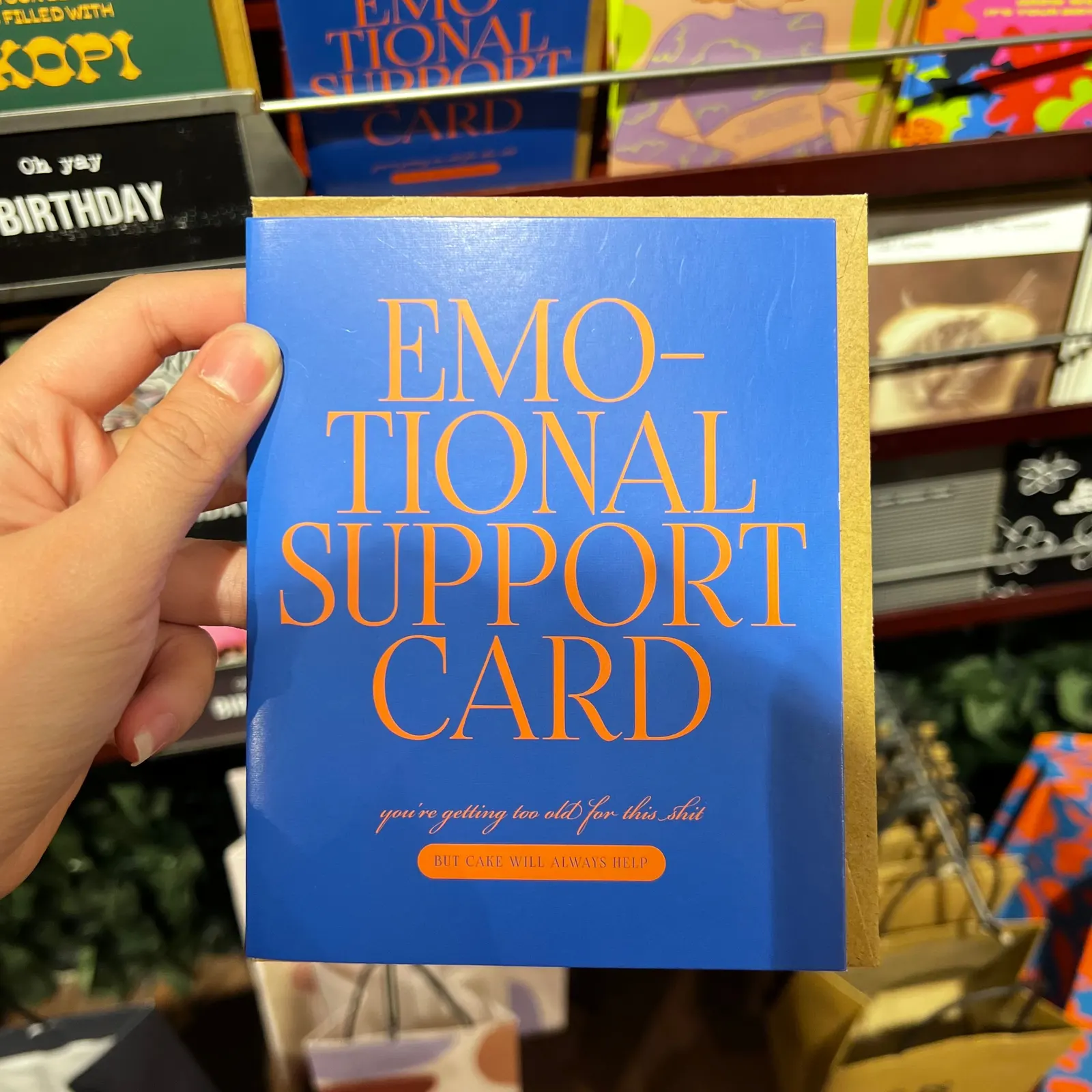 A hand holding a blue greeting card that reads “Emotional support card” in front of a shelf of colourful cards.