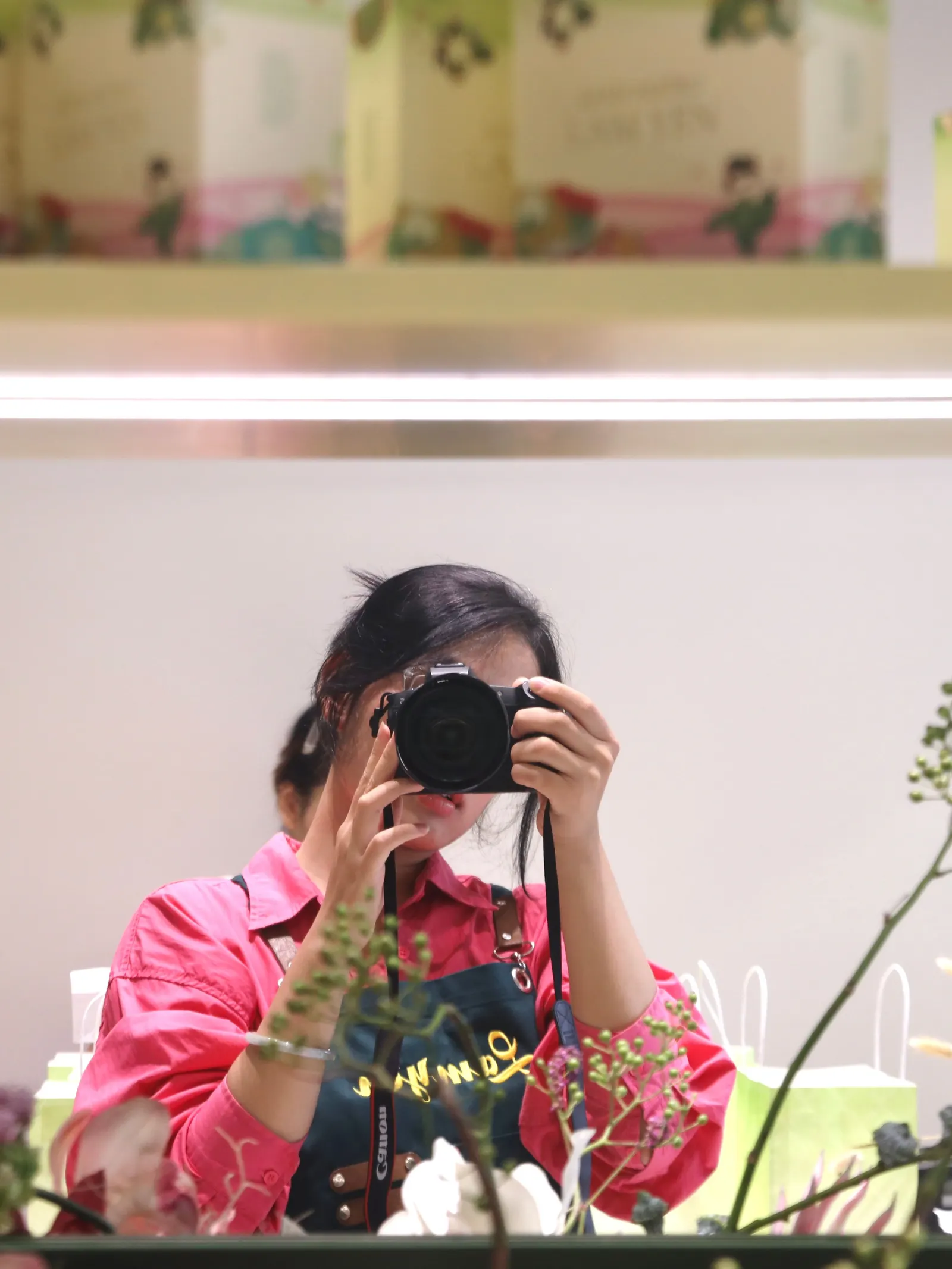Cam Anh in a pink shirt and apron photographing her reflection with a camera, between shelves of green gift boxes and sprigs of dried flowers.