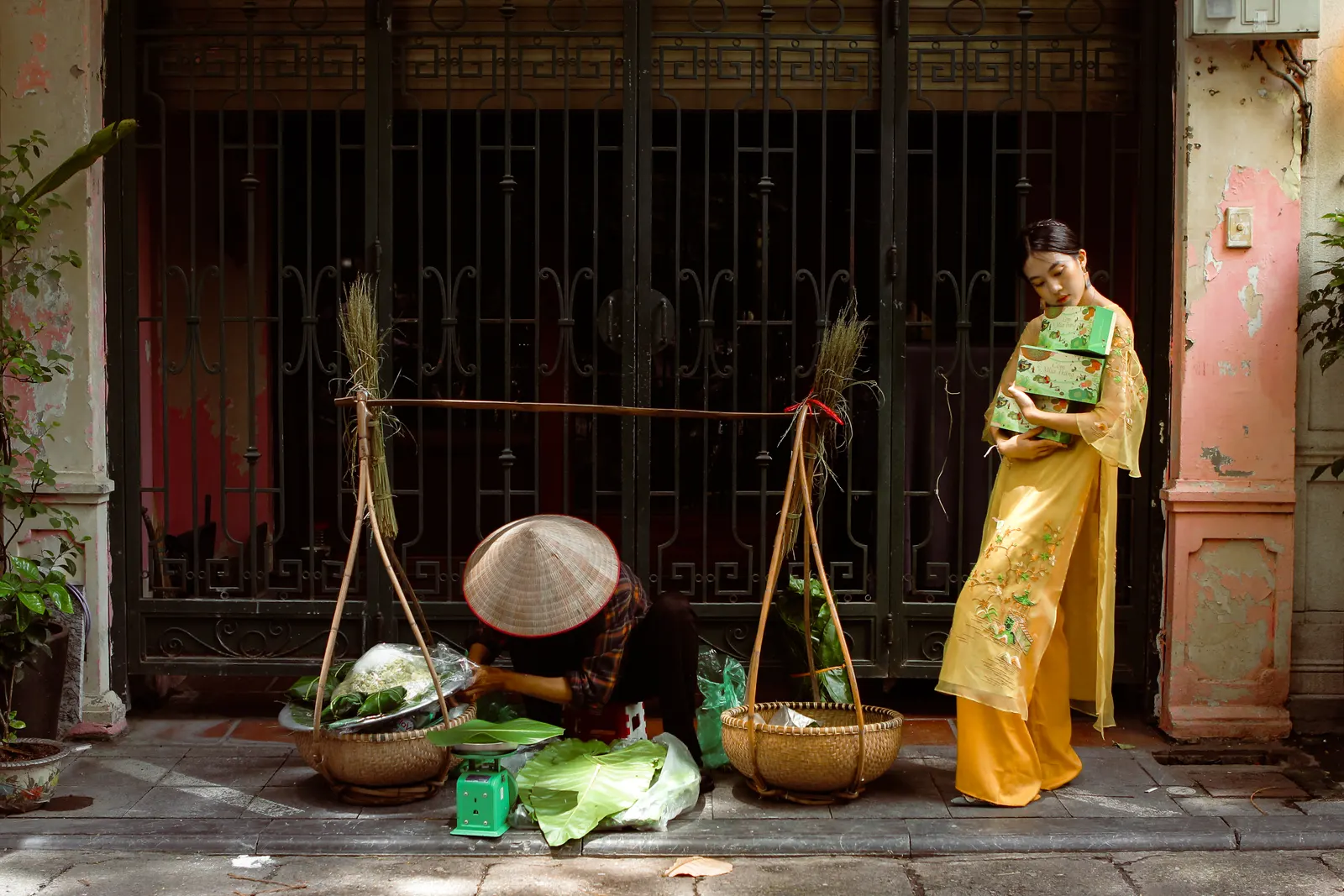A street vendor in a conical hat crouching between two shoulder-pole baskets of greens, with a woman in a yellow áo dài standing by a wrought-iron gate.