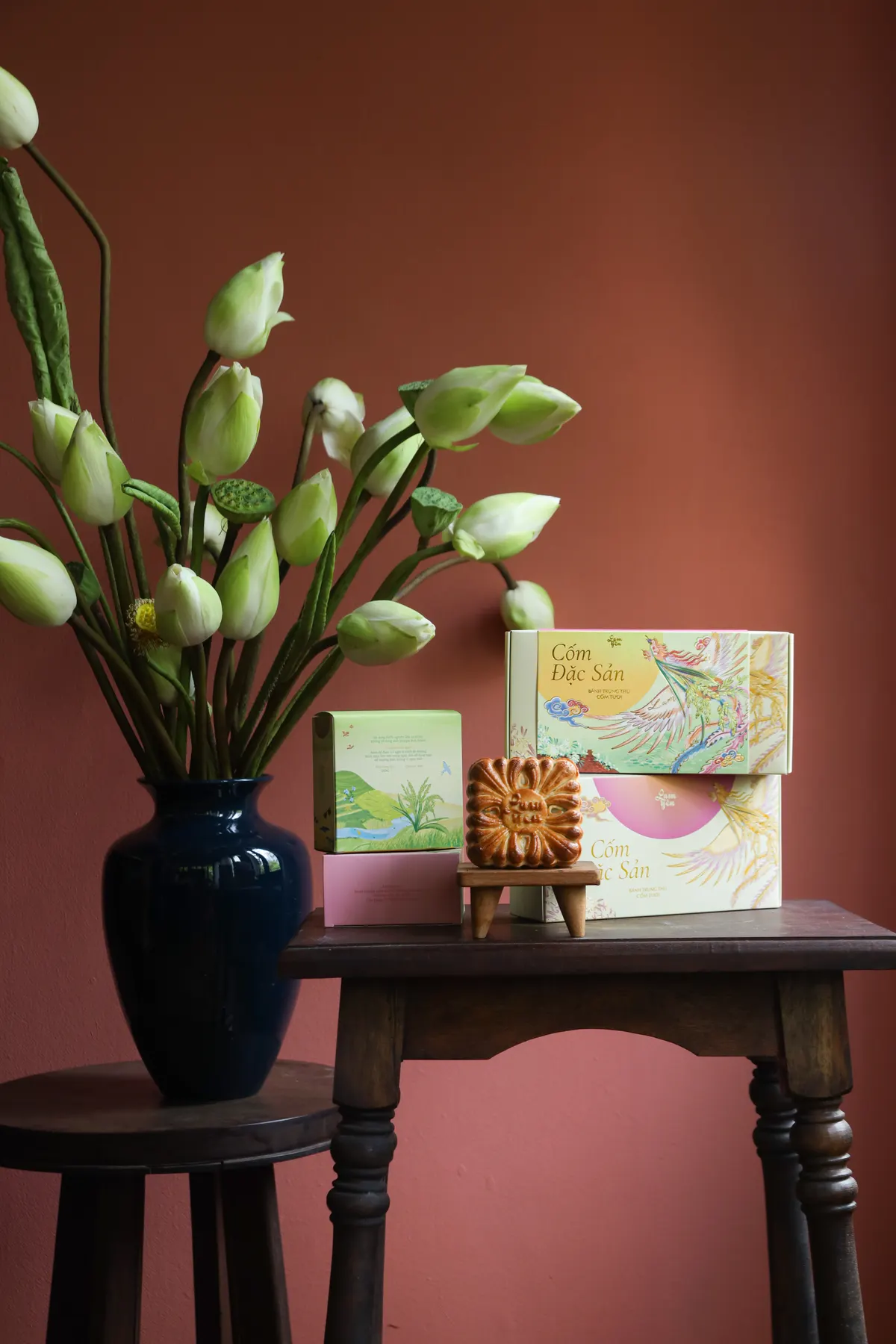 White lotus buds in a dark blue vase on a small table, with Cốm Đặc Sản gift boxes and a mooncake on a stand beside it, against a dusty-pink wall.