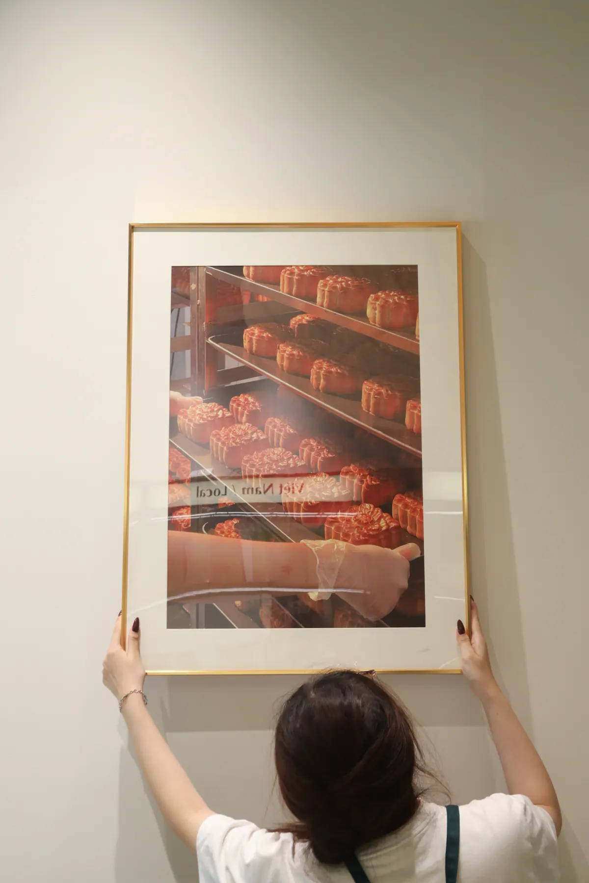 A woman holding up a framed photo of mooncakes on a rack against a pale wall.