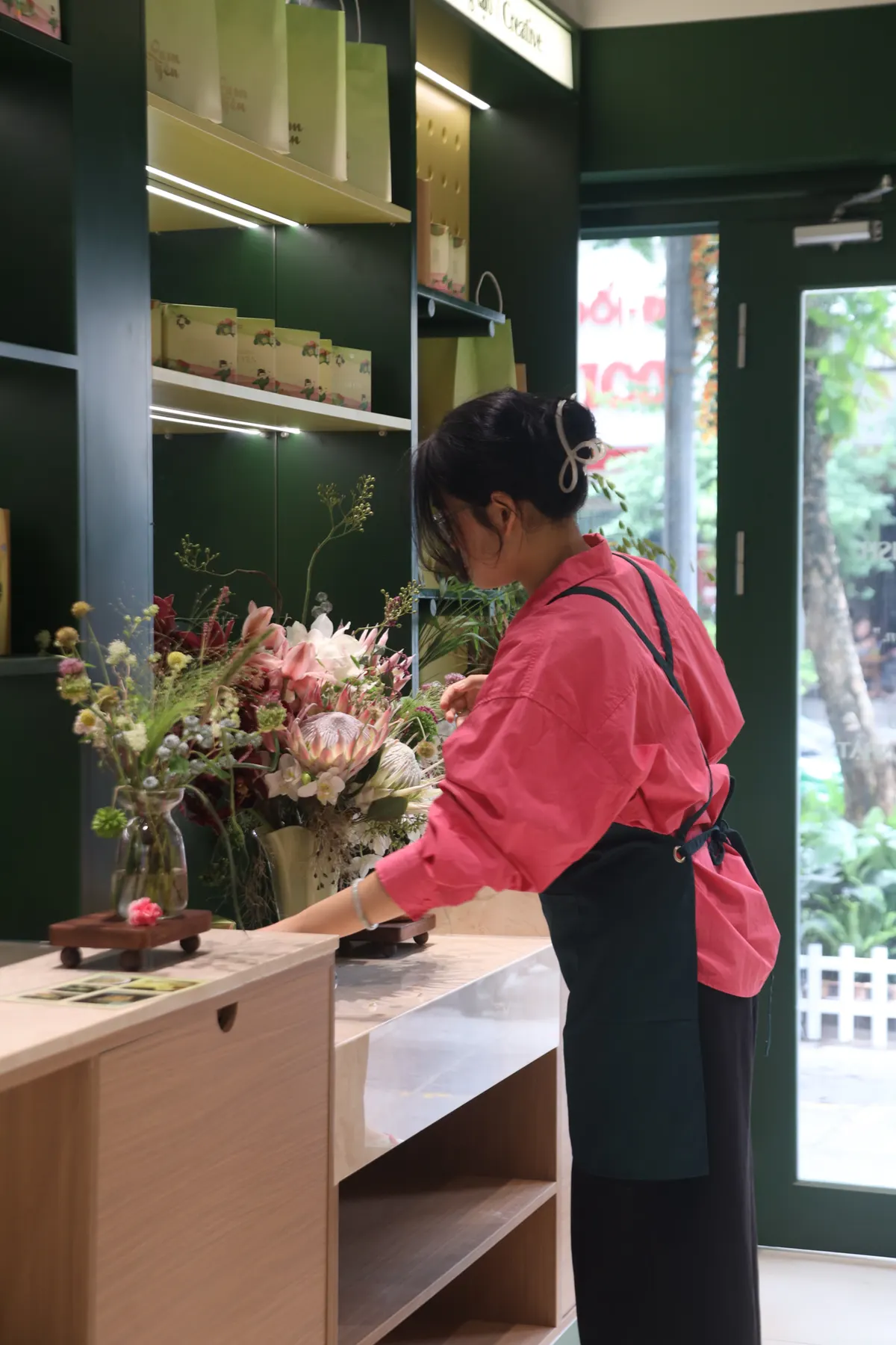 A shop assistant in a pink shirt arranging flowers on a wooden counter in the green shop.