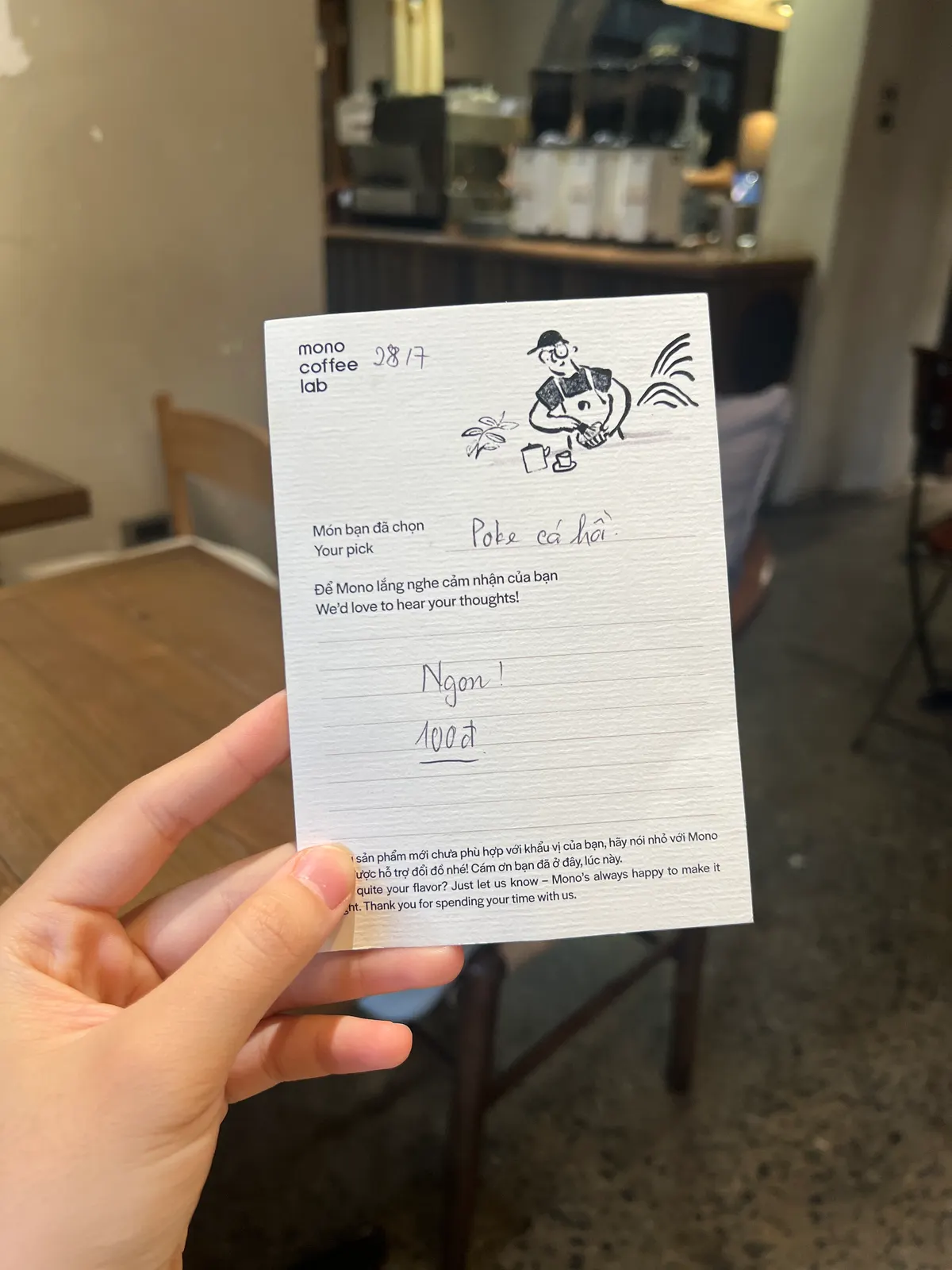 A hand holding up a Mono Coffee Lab guest opinion card, filled in by hand, in the café (card 4 of 9).