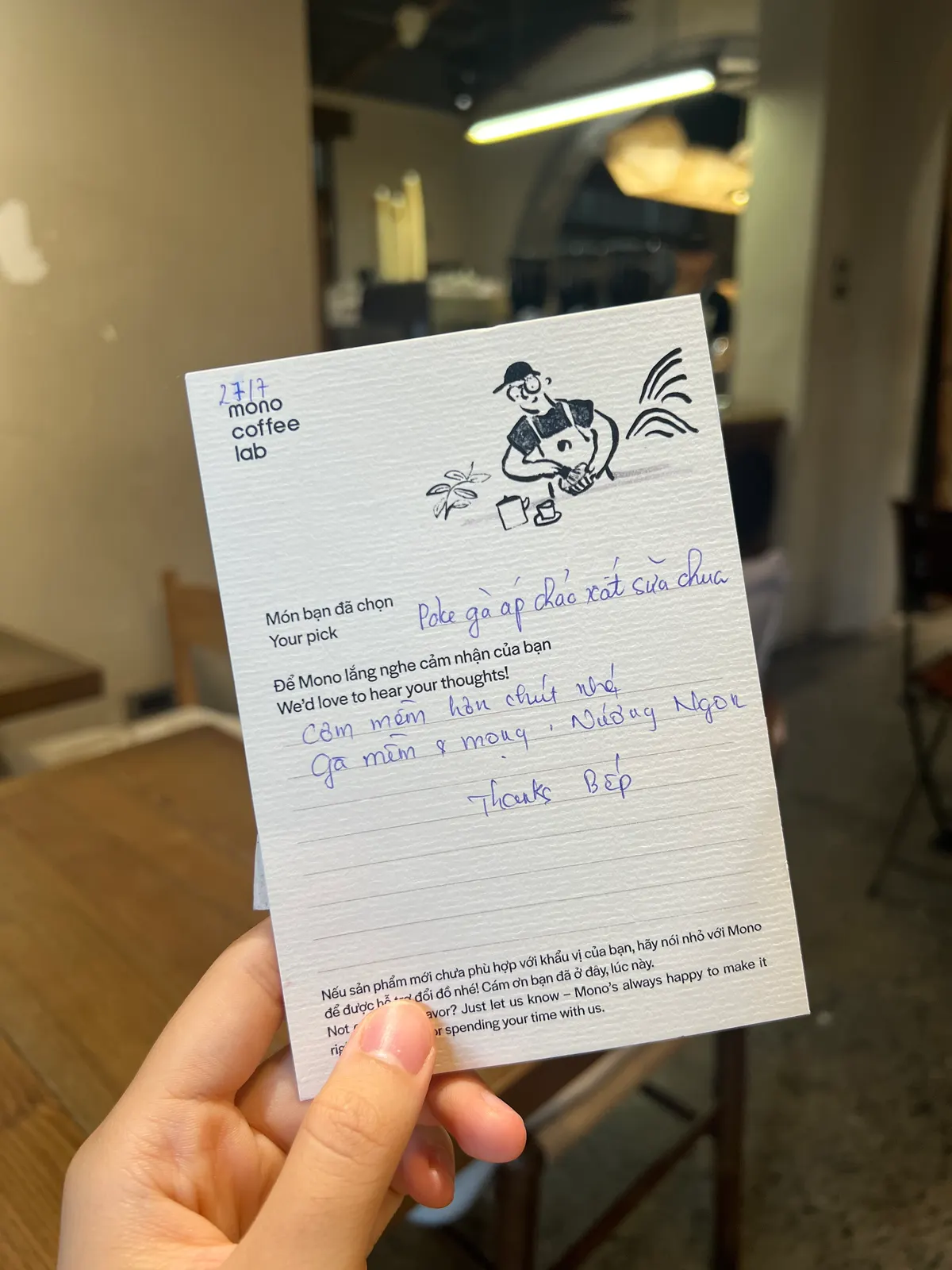 A hand holding up a Mono Coffee Lab guest opinion card, filled in by hand, in the café (card 8 of 9).