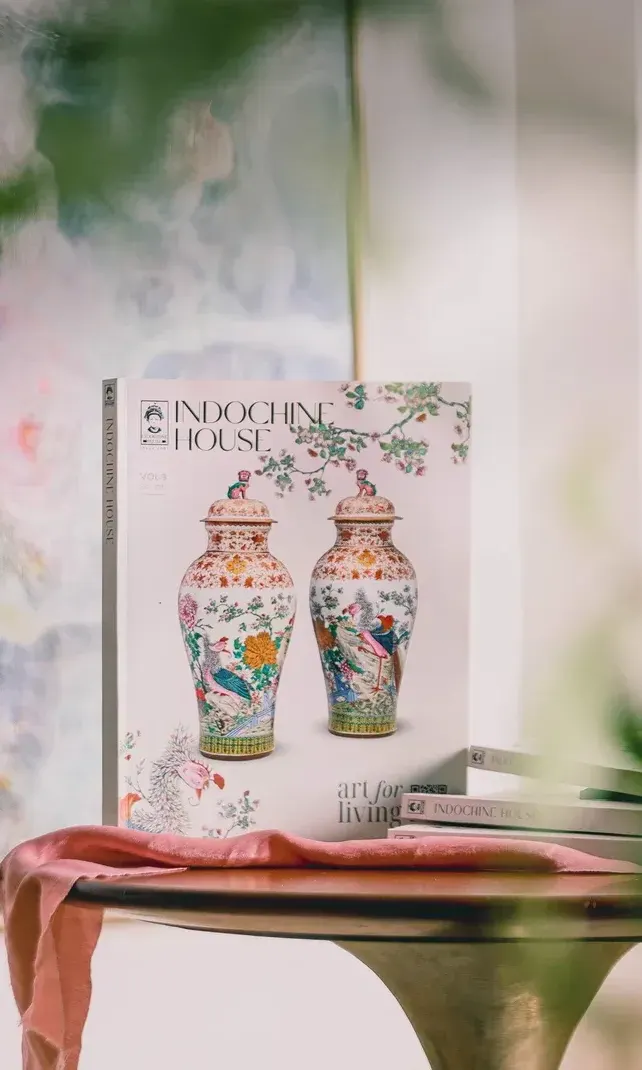 The Indochine House catalogue standing on a round table in front of a soft abstract painting.