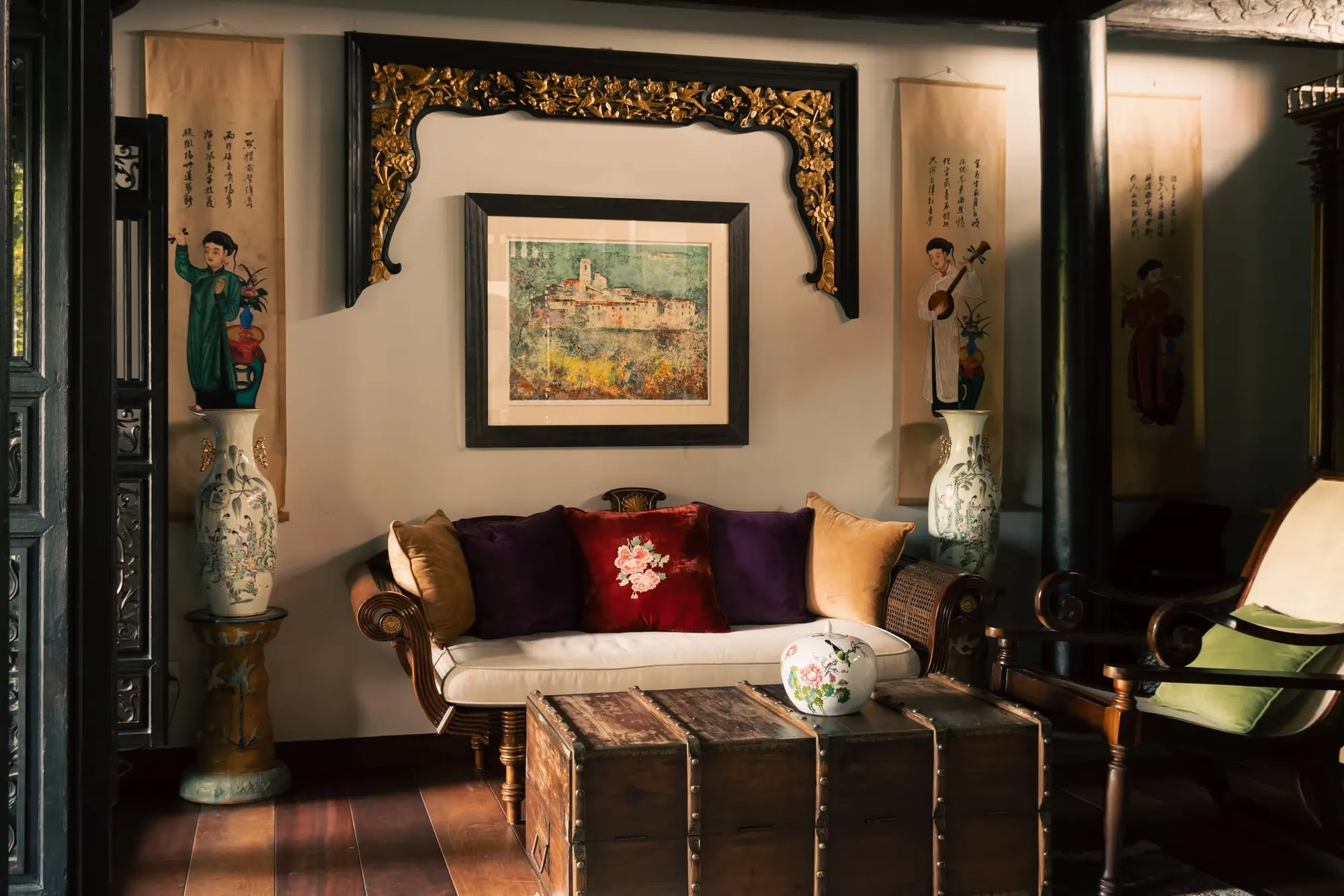 A dark-wood Indochine villa sitting room in warm light: a daybed with plum, red and gold cushions under a framed landscape and a carved gilded lintel, tall painted vases and hanging scrolls on both sides, a leather-bound chest in front.
