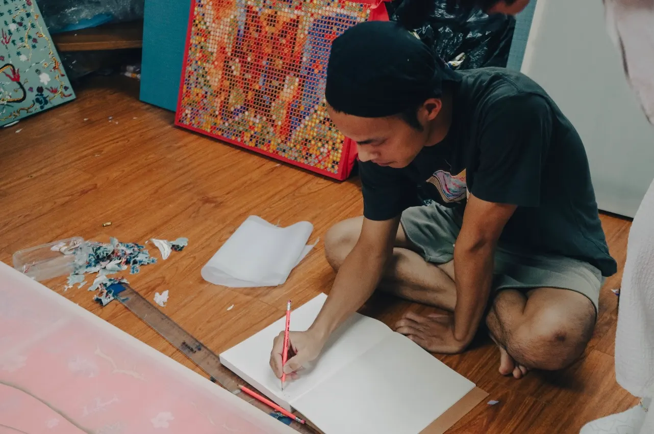 An artist in a bandana sitting on a wooden floor, drawing in a notebook, with paintings behind him.