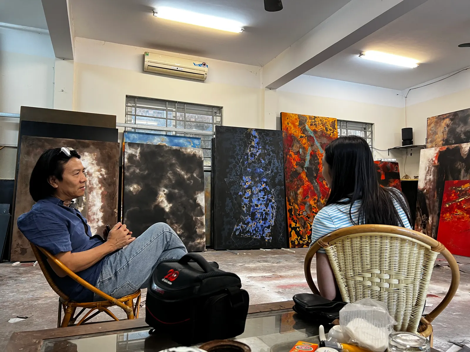 An artist and a visitor sitting in a studio with large dark and orange canvases leaning behind them.