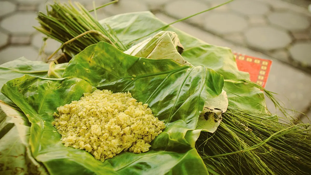 A small heap of young green rice (cốm) on a taro leaf, with bundles of grass beside it on a paved street.