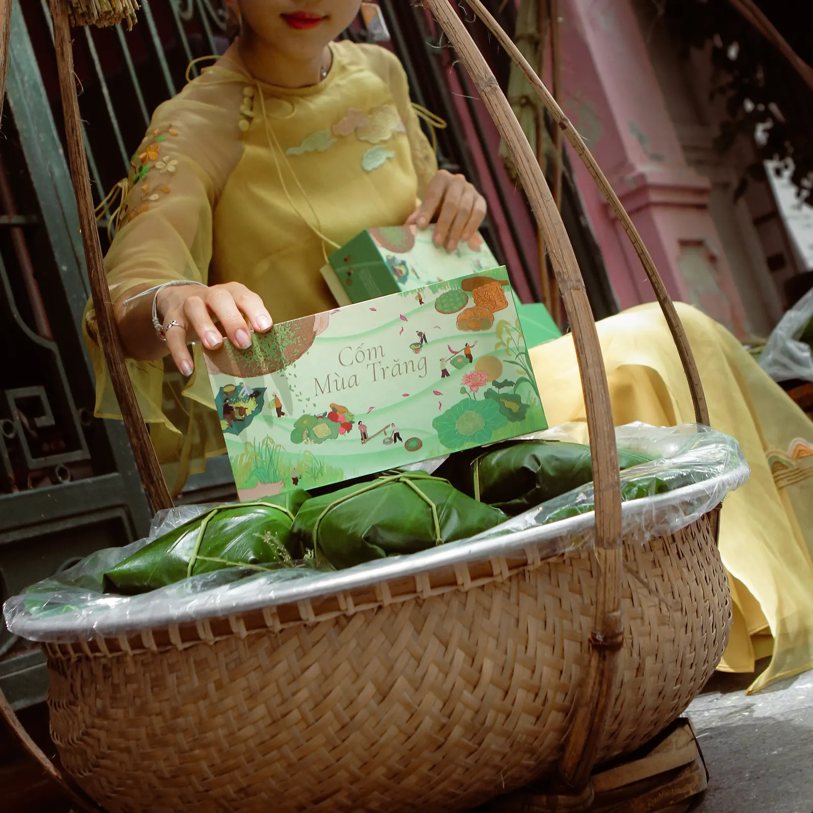 A woman in a yellow ao dai holding a green Cốm Mùa Trăng gift box above a bamboo basket of leaf-wrapped cakes.
