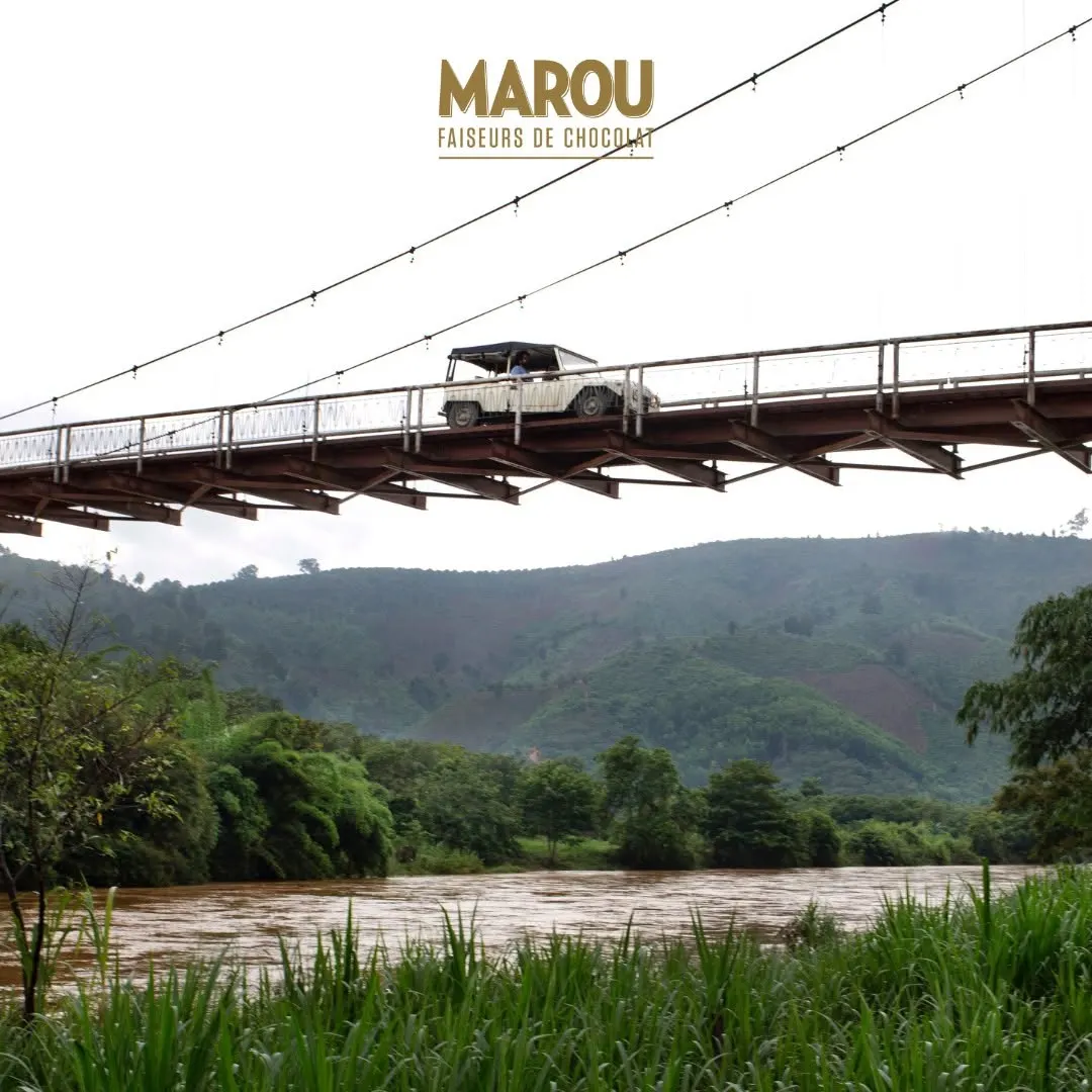 A white jeep crossing a long suspension bridge over a river valley, green hills behind.