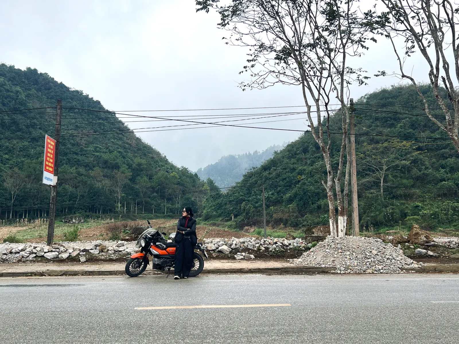 Cam Anh in a black jacket and sunglasses standing beside an orange motorbike on a mountain road, green hills behind.